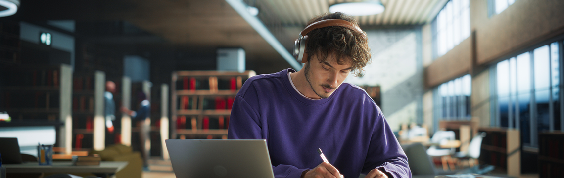 Young man with headphones working on laptop and taking notes